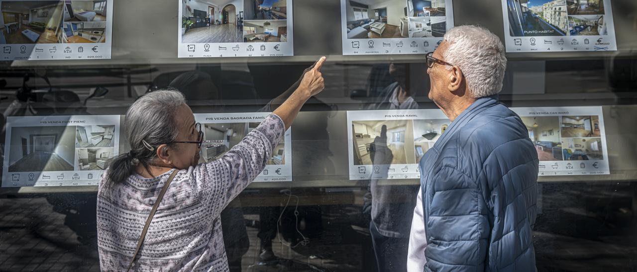 Dos personas miran carteles de pisos de alquiler y compra en una inmobiliaria de Barcelona.